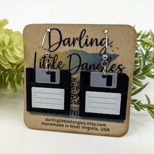 May include: Black and white floppy disk earrings with silver hooks. The earrings are displayed on a brown card with the text "Darling Little Dangles" and the Etsy shop URL. The earrings are a unique accessory.