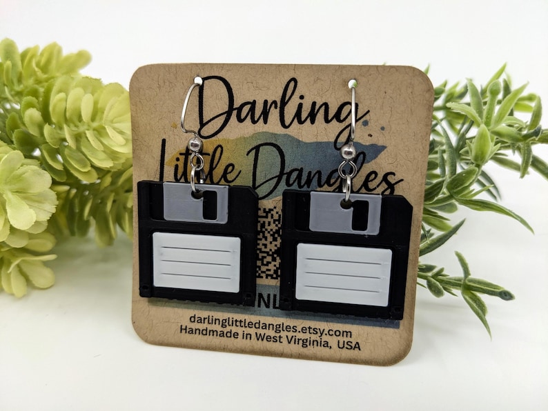 May include: Black and white floppy disk earrings with silver hooks. The earrings are displayed on a brown card with the text "Darling Little Dangles" and the Etsy shop URL. The earrings are a unique accessory.