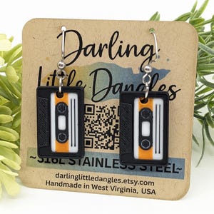 May include: Black and white cassette tape earrings with orange accents, hanging from silver hooks. The earrings are displayed on a brown card with the text "Darling Little Dangles" and the shop's website.