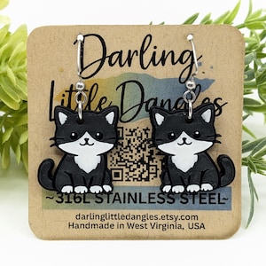 May include: A pair of black and white cat earrings with stainless steel hooks. The earrings are on a brown card with the text "Darling Little Dangles" and the website address. The earrings feature a cute cat design.