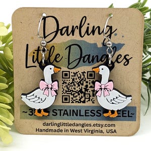 May include: A pair of dangle earrings featuring white geese with pink bows, orange beaks and feet, and black accents. The earrings are on a brown card with the text "Darling Little Dangles" and "Stainless Steel".