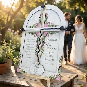 Op de afbeelding: Een wit houten trouwteken met een marmerachtige achtergrond, bloemaccenten en tekst. Het bord bevat een gevlochten touwdetail en een label met namen en een datum. Op de achtergrond loopt een stel.