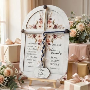 May include: A white wooden wedding sign with floral accents and a braided rope design. The sign features the text "They are no longer two..." and "A cord of three strands is not easily broken." Includes the names Adam & Sophia.