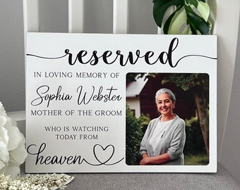 Könnte beinhalten: Weißer Holzrahmen mit einem Foto einer Frau und dem Text "reserviert" in schwarzer Kursivschrift. Der Text unter dem Foto lautet "In liebevoller Erinnerung an Sophia Webster, Mutter des Bräutigams, die heute vom Himmel aus zuschaut".