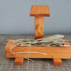 Könnte beinhalten: Eine hölzerne Krippe mit einem mit Heu gefüllten Trog und einem kleinen Holzdach. Die Krippe ist orange lackiert und hat einen kleinen Holzgriff an der Seite.