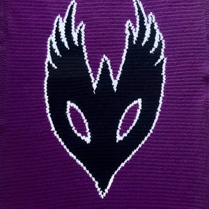 May include: A crocheted blanket in shades of purple, featuring a central black mask design with white outlining. The mask has a heart-shaped base and stylized wings. White decorative elements are at the top and bottom.
