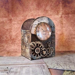 May include: Steampunk desk tidy featuring a clock face, gears, and rivets. The clock has Roman numerals and a vintage aesthetic. The organiser is a mix of metallic bronze and dark brown, with a distressed finish. The design incorporates a lightbulb and map details.