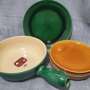 Op de afbeelding: Een verzameling keramische serveerschalen in groen, geel en oranje. De set bevat een fonduepan met een handvat, een grote groene schaal en een stapel kleinere borden. De borden hebben reliëfontwerpen van eten en drinken.