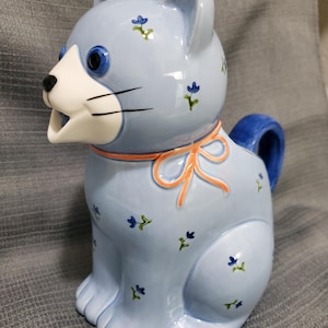 Puede incluir: Una tetera de cerámica azul claro con forma de gato, con hocico blanco y ojos azules. La tetera está decorada con pequeñas flores azules y un lazo color melocotón alrededor del cuello. El asa y la cola son azules.