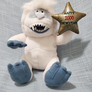 May include: A plush white abominable snowman toy with blue accents, holding a gold star-shaped ornament. The ornament has the words "HAPPY 2000 NEW YEAR" in red and green. The toy has a toothy grin and blue feet.