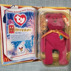 Puede incluir: Un oso de peluche Millennium the Bear en una funda de plástico transparente, con cuerpo rojo y cinta dorada. El embalaje incluye un folleto con el texto "Ronald McDonald House Charities" y "Millennium the Bear."