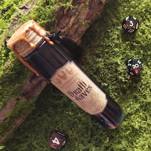 May include: A dark glass potion bottle with a cork stopper sealed with orange wax. The label reads "Death Saves" with skull graphics. Several dice are scattered on a green surface, suggesting a tabletop game theme.