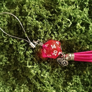 Könnte beinhalten: Ein Schlüsselanhänger mit einem transparenten roten zwanzigseitigen Würfel mit weißen Zahlen, einem silberfarbenen Metallherz-Anhänger mit filigranem Muster und einer leuchtend pinken Quaste, befestigt an einer dünnen silbernen Kordelschlaufe.
