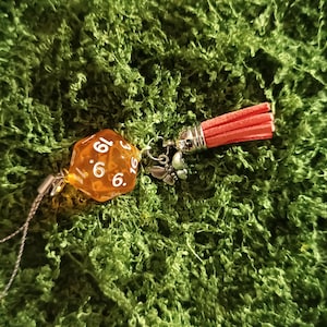 Può includere: Un dado a venti facce arancione traslucido con numeri bianchi, attaccato a un anello in metallo argentato con un piccolo ciondolo a forma di farfalla e una nappa in camoscio color corallo, collegato a un sottile cordino grigio.