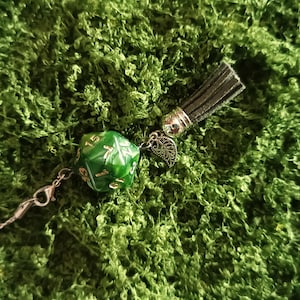 Puede incluir: Un dado de veinte caras verde con números dorados unido a un cierre de mosquetón de metal plateado, un pequeño dije de hoja plateada y una borla de ante gris oscuro. El dado presenta un acabado jaspeado y una fina cadena de metal plateado.