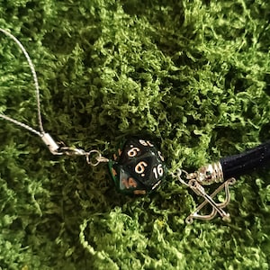 May include: A black twenty-sided die with gold numbers attached to a silver-toned metal loop. The charm features a small silver-toned bow and arrow pendant and a short dark blue fabric tassel, all connected by a thin silver-toned metal chain.