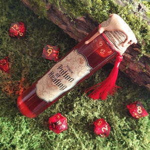 May include: A glass potion bottle filled with a red liquid, labeled "Minor Potion of Healing." The bottle is sealed with a cork and twine, adorned with a red tassel. Scattered around are several red polyhedral dice, suggesting a tabletop gaming theme.
