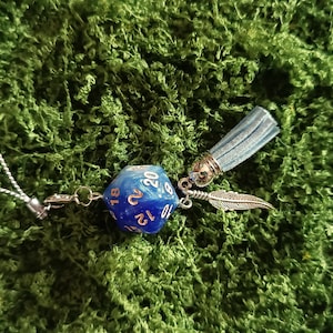 Può includere: Un ciondolo a forma di dado a venti facce blu e bianco con numeri dorati, attaccato a un cordino argentato. Il ciondolo include una nappa azzurra e un ciondolo a forma di piuma in metallo argentato, collegati da piccoli anelli in metallo.