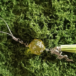 Puede incluir: Un colgante para teléfono que presenta un dado de veinte caras amarillo translúcido con números blancos, un dije de metal plateado con forma de serpiente y una borla de piel sintética verde claro, unidos a un fino cordón plateado.