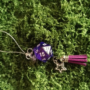 Pode incluir: Um pingente de dado de vinte lados roxo com números brancos, preso a um laço de cordão prateado. O pingente inclui um pequeno pendente de estrela prateado e uma borla curta de camurça roxo-escura com uma tampa metálica prateada.