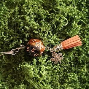 Può includere: Un ciondolo per telefono con un dado a venti facce in gradazione nero e rame, un ciondolo a forma di foglia d'acero color argento e una nappa in camoscio marrone chiaro. I componenti sono collegati da anellini in metallo argentato a un cordino.