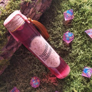 Könnte beinhalten: Eine Glasflasche mit einer tiefroten Flüssigkeit, beschriftet mit "Feywild Essence", ist mit Bindfaden und rosa Wachs versiegelt. Eine kleine goldene Quaste hängt oben. Mehrere schillernde, vielseitige Würfel liegen auf grünem Moos verstreut.