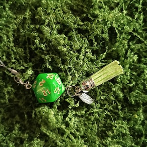 Puede incluir: Un dije de dado de veinte caras verde con números dorados, unido a un cordón plateado. El dije incluye una borla de aspecto de gamuza verde claro y un pequeño colgante de hoja de metal plateado conectados por una argolla plateada.