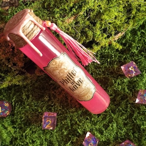 May include: A pink potion bottle with a label reading "Philter of Love" and a pink tassel, resting on green moss. Several dice with purple and gold accents are scattered around the bottle, creating a fantasy theme.