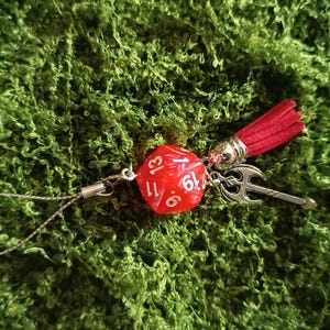 Puede incluir: Un dije de dado de veinte caras rojo con números blancos, unido a un lazo de metal plateado. El dije incluye una pequeña borla de ante rojo y un colgante de hacha de metal plateado, conectados por un cordón plateado fino para colgar.