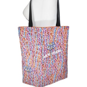 May include: A tote bag with black straps and a colorful design featuring a repeated pattern of Pixy Stix candy. The bag is made of canvas and has a white background with orange, blue, and purple Pixy Stix.
