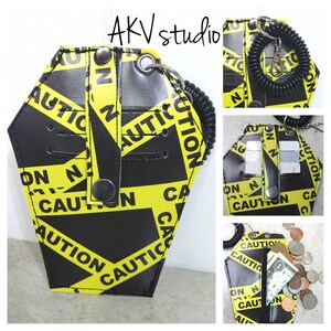 May include: A black and yellow leather wallet with a caution tape pattern. The wallet has a snap closure and a key ring. It is designed to look like a coffin.