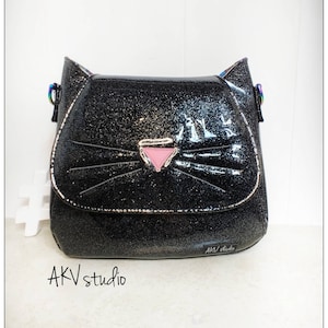 May include: A black glittery cat-shaped crossbody bag with a pink nose and silver trim. The bag has a flap closure and rainbow-colored strap attachments. The text "AKV studio" is printed on the bottom right of the bag.
