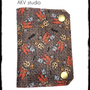 May include: A brown wallet with a repeating pattern of orange and gray scarecrow figures, black bats, and jack-o'-lanterns. The wallet is secured with two gold-colored snaps. The text "AKV studio" and "shop@AKVstudio.etsy.com" are also visible.