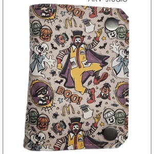 May include: A beige wallet with a Halloween-themed pattern featuring Ronald McDonald, ghosts, and the McDonald's logo. The wallet has a snap closure and the text "BOO!" is visible. The text "AKV studio" and "AKVstudio.etsy.com" are also present.