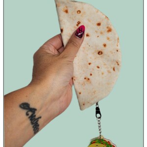 May include: A light-colored tortilla-shaped pouch with a zipper, held by a hand with a tattoo. Attached is a taco-shaped keychain with the text "It's okay to fall apart sometimes. Tacos fall apart and we still love them."