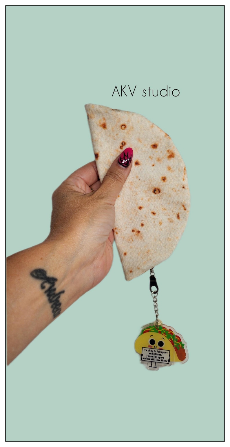 May include: A light-colored tortilla-shaped pouch with a zipper, held by a hand with a tattoo. Attached is a taco-shaped keychain with the text "It's okay to fall apart sometimes. Tacos fall apart and we still love them."