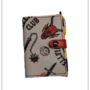 May include: A white wallet with a black and red design featuring a dice, a chain with a spiked ball, a sword, and a demon head. The wallet has a red strap with a black button.