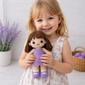 Può includere: Una bambola all'uncinetto fatta a mano con capelli castani, un vestito viola e scarpe abbinate. La bambola ha un viso sorridente con guance rosee. La bambola è tenuta in braccio da un bambino sorridente. Lo sfondo include lavanda e un cestino di vimini.
