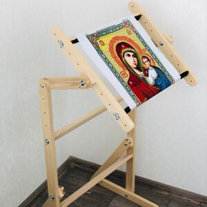 Op de afbeelding: Een houten borduurstandaard met een kantelbaar bovenblad en verstelbare hoogte. De standaard is gemaakt van natuurlijk hout en heeft een donkerbruine afwerking. Een borduurproject met kruissteek is op de standaard te zien.