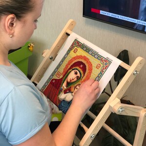 Op de afbeelding: Een persoon werkt aan een diamant schilderij van een religieus icoon. Het schilderij staat op een houten standaard. Het icoon is van de Maagd Maria en kind. Het schilderij is omgeven door een gele en oranje rand. De tekst op het schilderij luidt "Слава Святой Богородице".