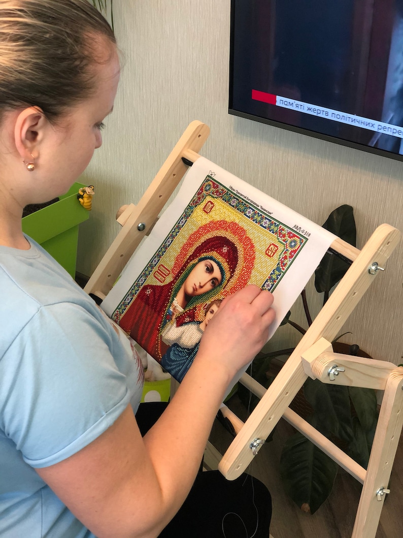 Op de afbeelding: Een persoon werkt aan een diamant schilderij van een religieus icoon. Het schilderij staat op een houten standaard. Het icoon is van de Maagd Maria en kind. Het schilderij is omgeven door een gele en oranje rand. De tekst op het schilderij luidt "Слава Святой Богородице".