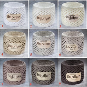 Könnte beinhalten: Neun Spulen mit Garn in verschiedenen Farben, darunter Weiß, Beige und Braun. Jede Spule hat ein Etikett mit der Aufschrift "Bobilon" und einem Farbnamen darunter.