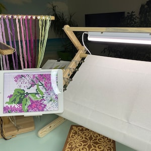 Puede incluir: Un bastidor de punto de cruz de madera con una tela blanca estirada sobre él. Una tableta que muestra un patrón de punto de cruz está a la izquierda del bastidor. Una barra de luz está unida a la parte superior del bastidor. Hay varios carretes de hilo de bordar colgando de un estante de madera.