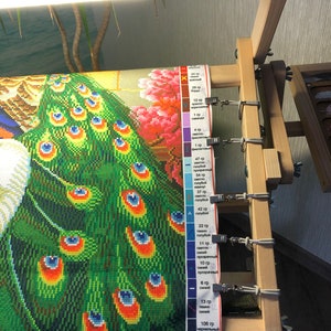 Op de afbeelding: Een close-up van een diamant schilderij van een pauw met een groene en blauwe staart. Het schilderij is op een houten frame met clips die het canvas op zijn plaats houden. Een kleurenkaart met nummers en beschrijvingen is zichtbaar aan de rechterkant van de afbeelding.