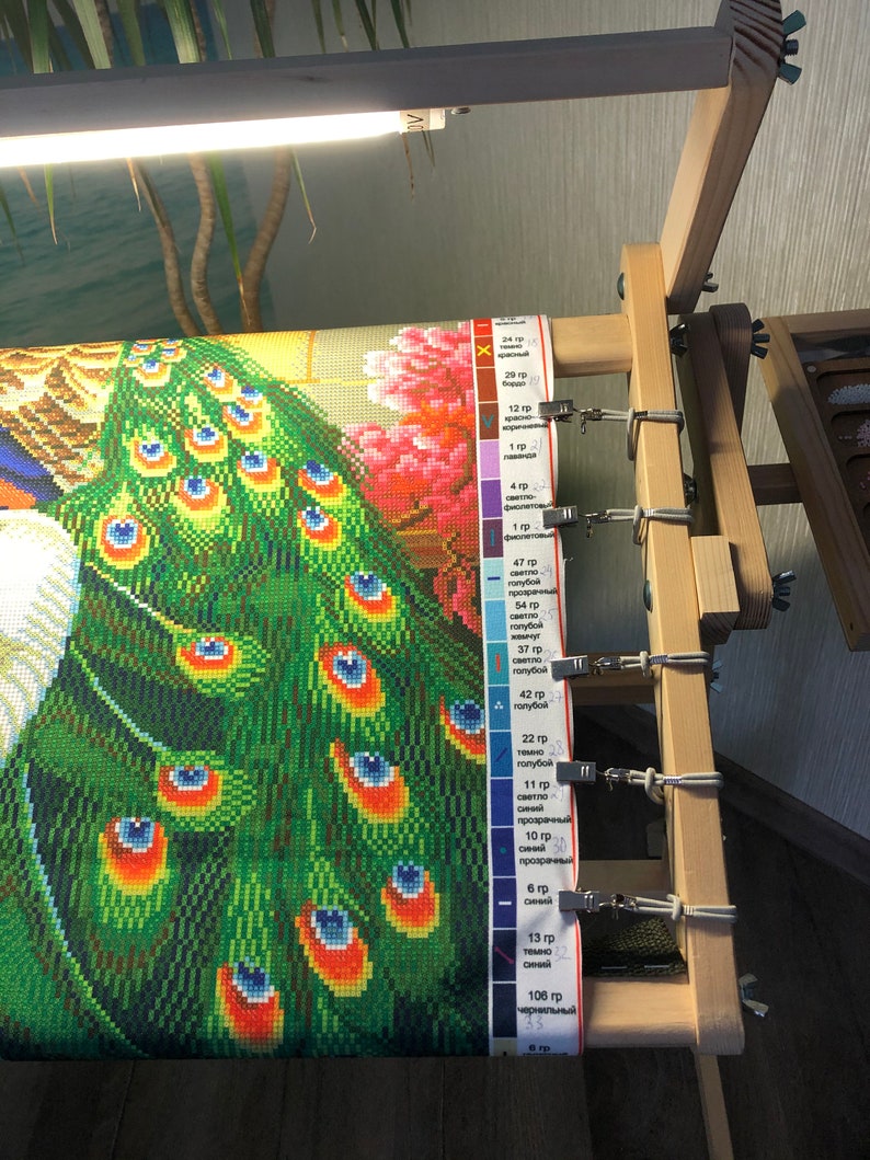Op de afbeelding: Een close-up van een diamant schilderij van een pauw met een groene en blauwe staart. Het schilderij is op een houten frame met clips die het canvas op zijn plaats houden. Een kleurenkaart met nummers en beschrijvingen is zichtbaar aan de rechterkant van de afbeelding.
