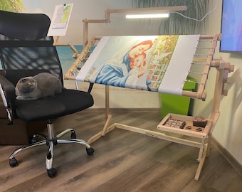 May include: A wooden embroidery stand with a light-colored wood finish. The stand has a built-in light and a tray for supplies. A canvas with a religious image is stretched on the stand. A gray cat sits on a black mesh office chair.