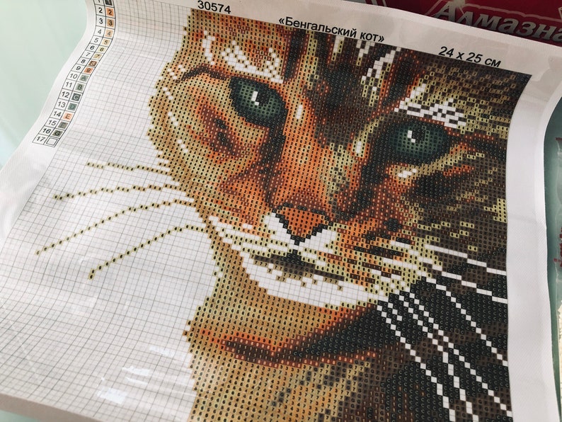 May include: Close-up of a diamond painting kit featuring a Bengal cat with green eyes. The kit is labeled "Bengal Cat" and measures 24 x 25 cm.