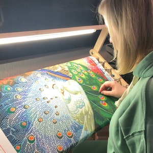 Pode incluir: Uma pessoa trabalhando em uma pintura de diamante de um pavão com um pássaro branco no centro. A pintura está em uma moldura de madeira com uma luz acima.