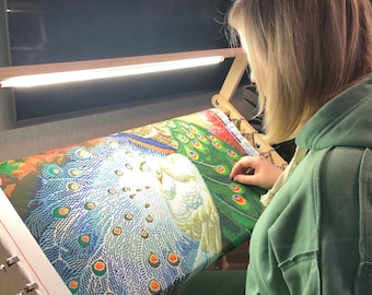May include: A person working on a diamond painting of a peacock with a white bird in the center. The painting is on a wooden frame with a light above it.