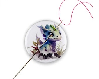 May include: A round button featuring a watercolour illustration of a small, blue and purple dragon with green accents. The dragon is surrounded by leaves and flowers. A needle and red thread are threaded through the button.